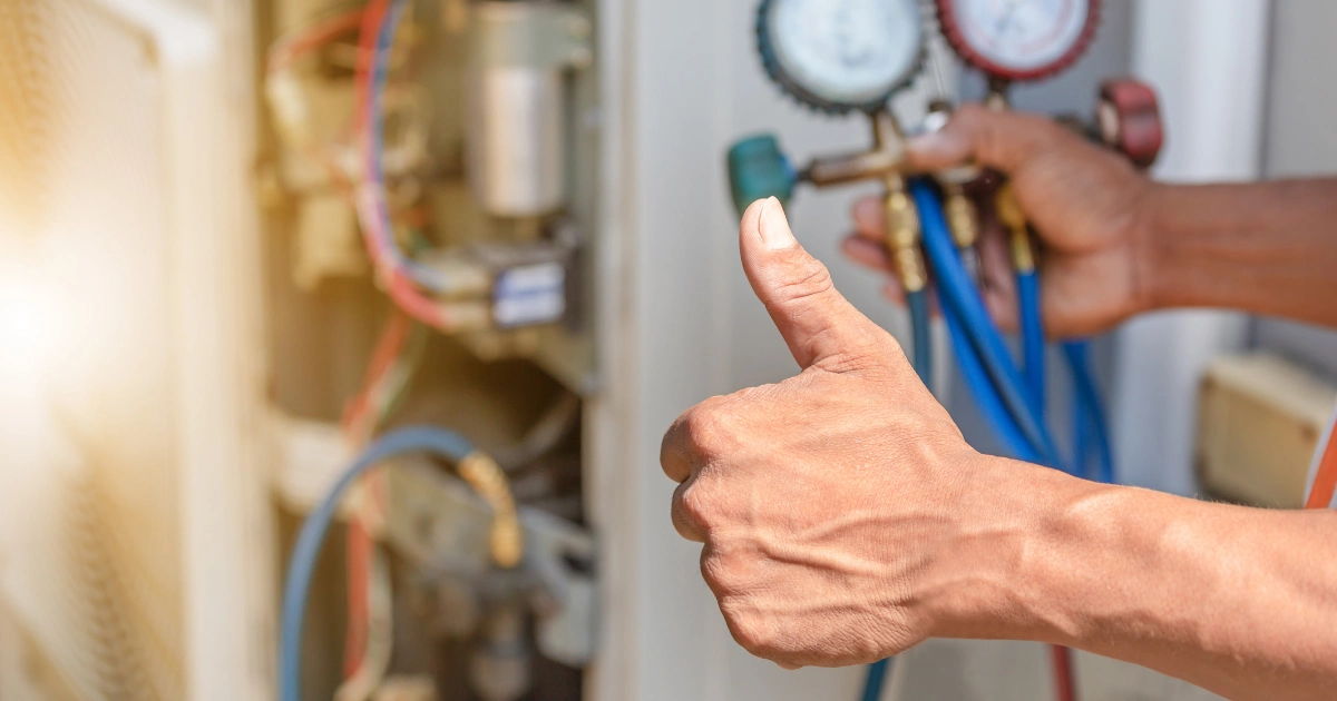 A person gives a thumbs up in front of HVAC equipment, holding gauges with hoses connected, indicating approval or satisfaction with the fall furnace tune-up and maintenance work being done.