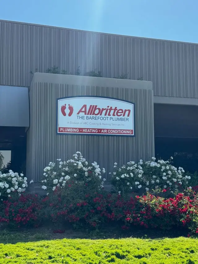 Exterior sign for Allbritten The Barefoot Plumber on a building with white and red flowering bushes in the foreground.