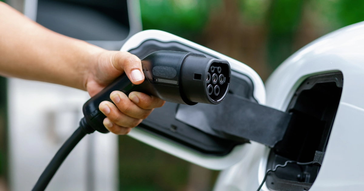 A person holds a charging plug and connects it to an electric vehicles charging port. The background features green, blurry foliage, and the vehicle is white.