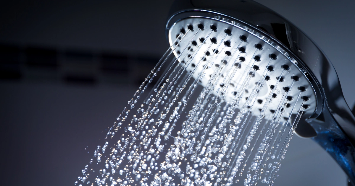 A close-up of a handheld showerhead spraying streams of water, with water droplets visible in mid-air against a blurred dark background.