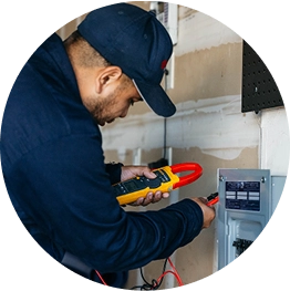 A person wearing a cap and blue jacket uses a digital clamp meter to check electrical wiring inside a control panel mounted on a wall in a garage or utility area.