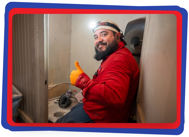 A man wearing a red shirt, orange gloves, and a headlamp sits in a small, partially constructed bathroom, giving a thumbs-up and smiling at the camera. There are construction tools and materials around him.