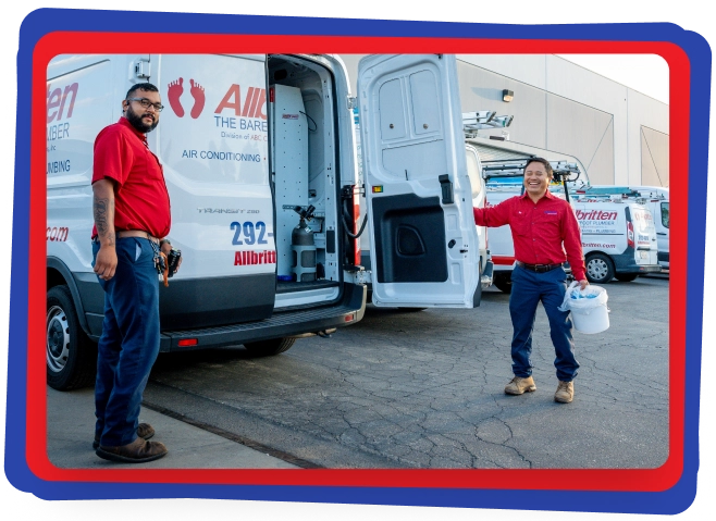 Two workers in red shirts and blue pants stand beside a white service van with its back doors open. One holds a white bucket. Several similar vans are parked nearby outside a commercial building.