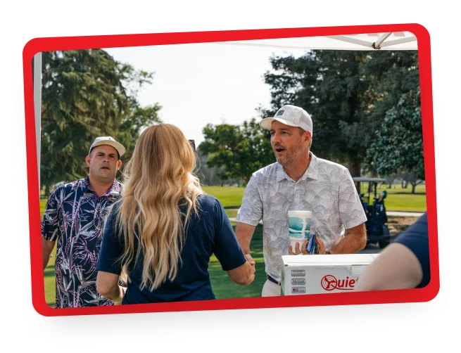 Three people converse outdoors at a golf course. A man in a patterned shirt and cap holds a drink and speaks to a woman with long blonde hair, while another man in a blue shirt and cap observes in the background.