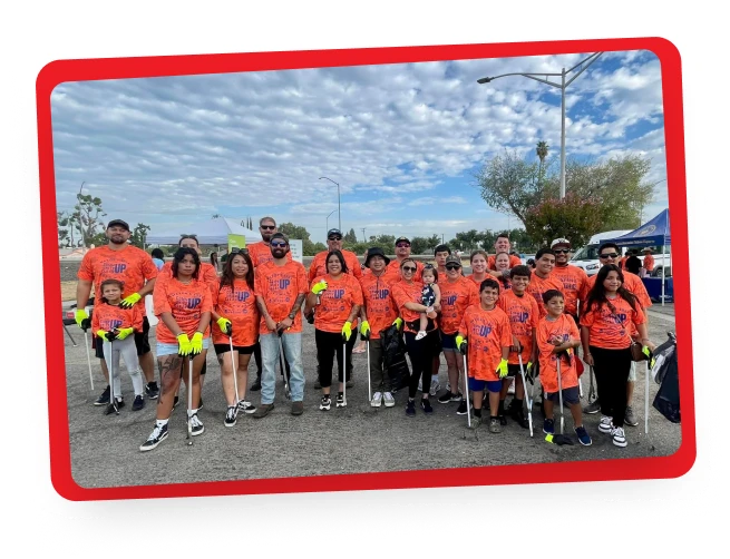 A group of people of various ages wearing matching orange shirts pose outdoors with litter pickers and trash bags, standing on pavement under a partly cloudy sky.