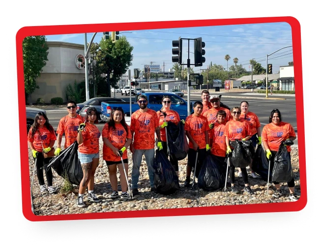 A group of people wearing matching orange shirts and yellow gloves stand on a rocky area by a street, holding large black garbage bags, participating in a community clean-up event.