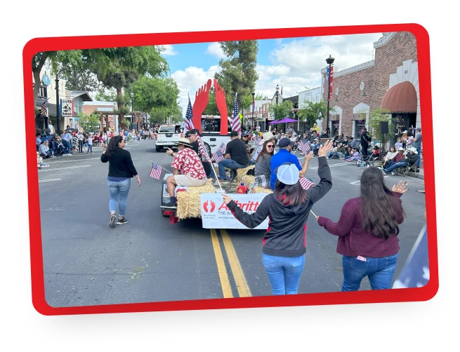 A parade scene shows people riding on and walking beside a float decorated with hay bales and American flags along a street lined with spectators and storefronts. Two women in the foreground wave toward the float.