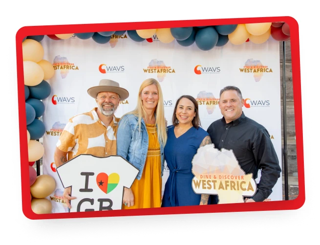 Four adults stand smiling in front of a backdrop with “WAVS” and “Dine & Discover West Africa” logos, holding themed signs. Decorative balloons frame the backdrop.