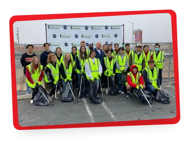 A group of people wearing yellow safety vests pose for a photo outdoors with trash bags and litter pickers. They stand in front of a banner and a chain-link fence along a paved area.