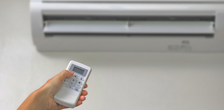 A hand holding a remote control is pointed at a wall-mounted air conditioning unit. The air conditioners vents are open and the display on the remote shows numbers.