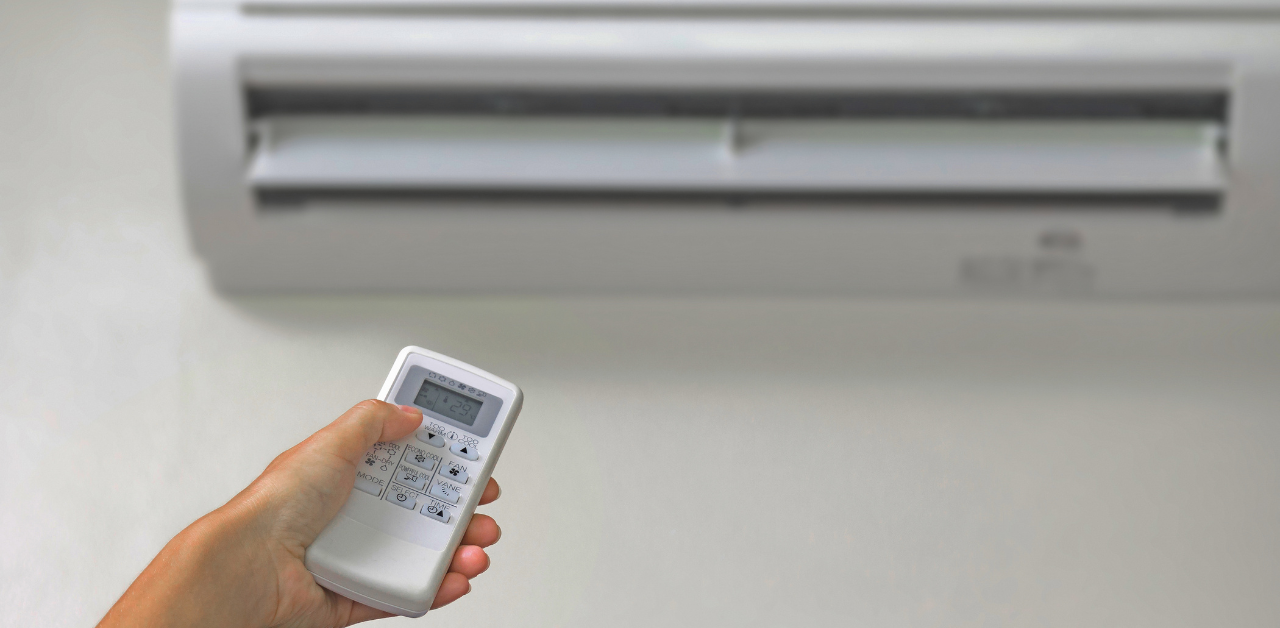 A hand holding a remote control is pointed at a wall-mounted air conditioning unit. The air conditioners vents are open and the display on the remote shows numbers.