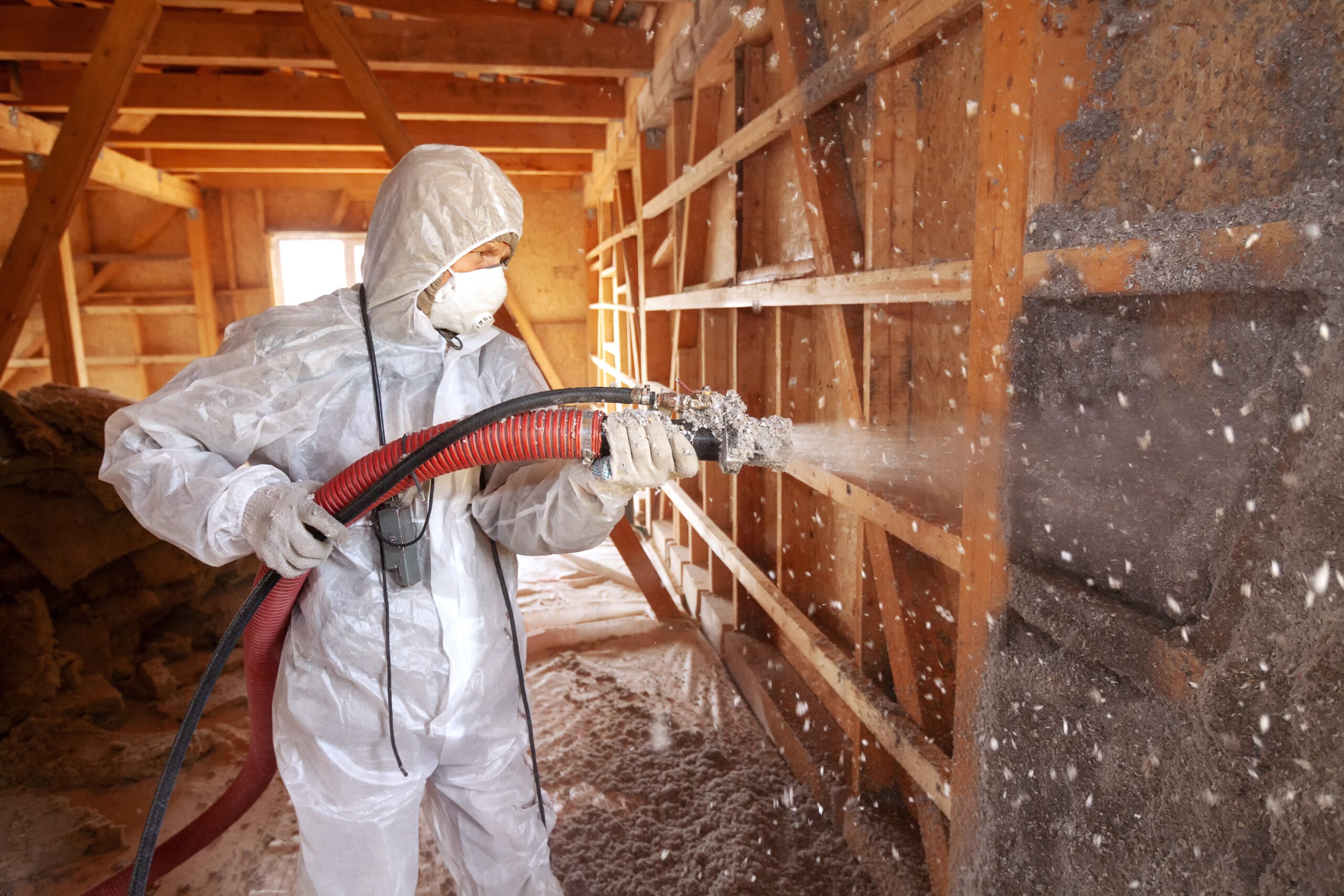 A person in protective clothing and a mask sprays insulation material onto a wall inside a wooden building under construction or renovation.