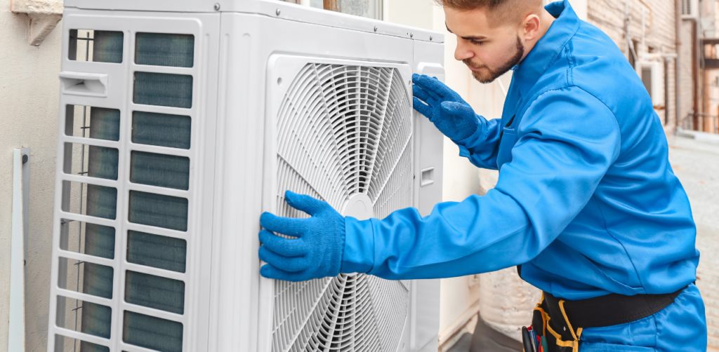 A technician in blue work clothes and gloves examines or installs an outdoor air conditioning unit attached to a building wall. Tools are visible on the technicians belt.