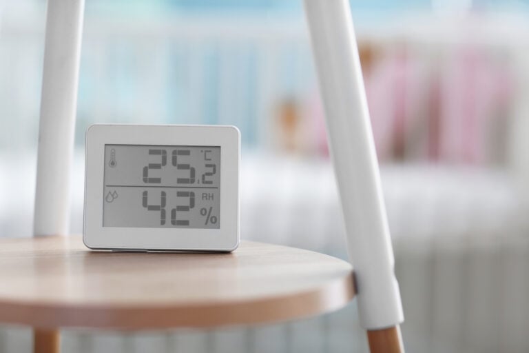 A digital thermometer and hygrometer sits on a wooden stool, displaying a temperature of 25.2°C and a humidity level of 42%. A blurred crib and blue wall are visible in the background.