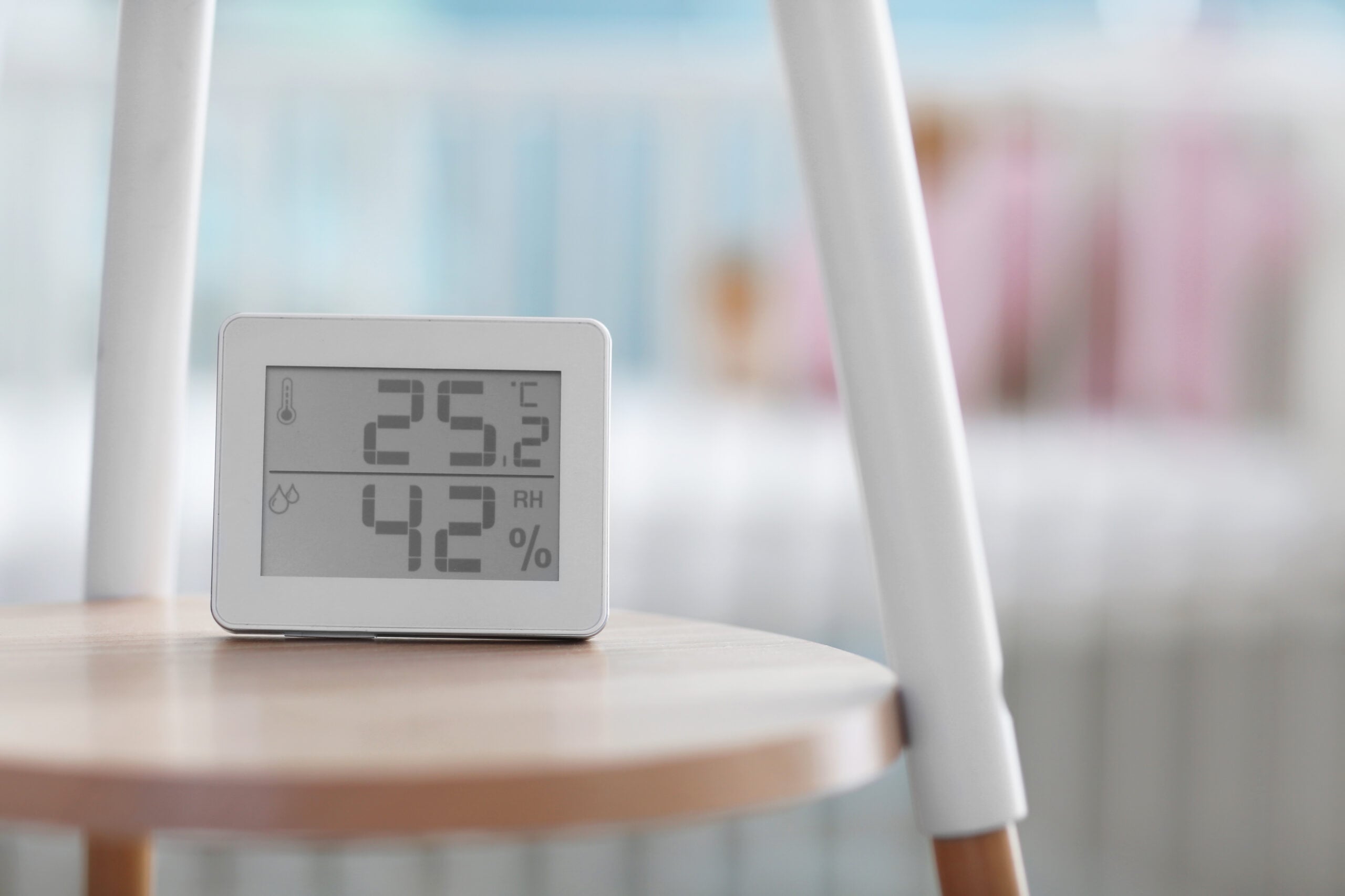 A digital thermometer and hygrometer sits on a wooden stool, displaying a temperature of 25.2°C and a humidity level of 42%. A blurred crib and blue wall are visible in the background.