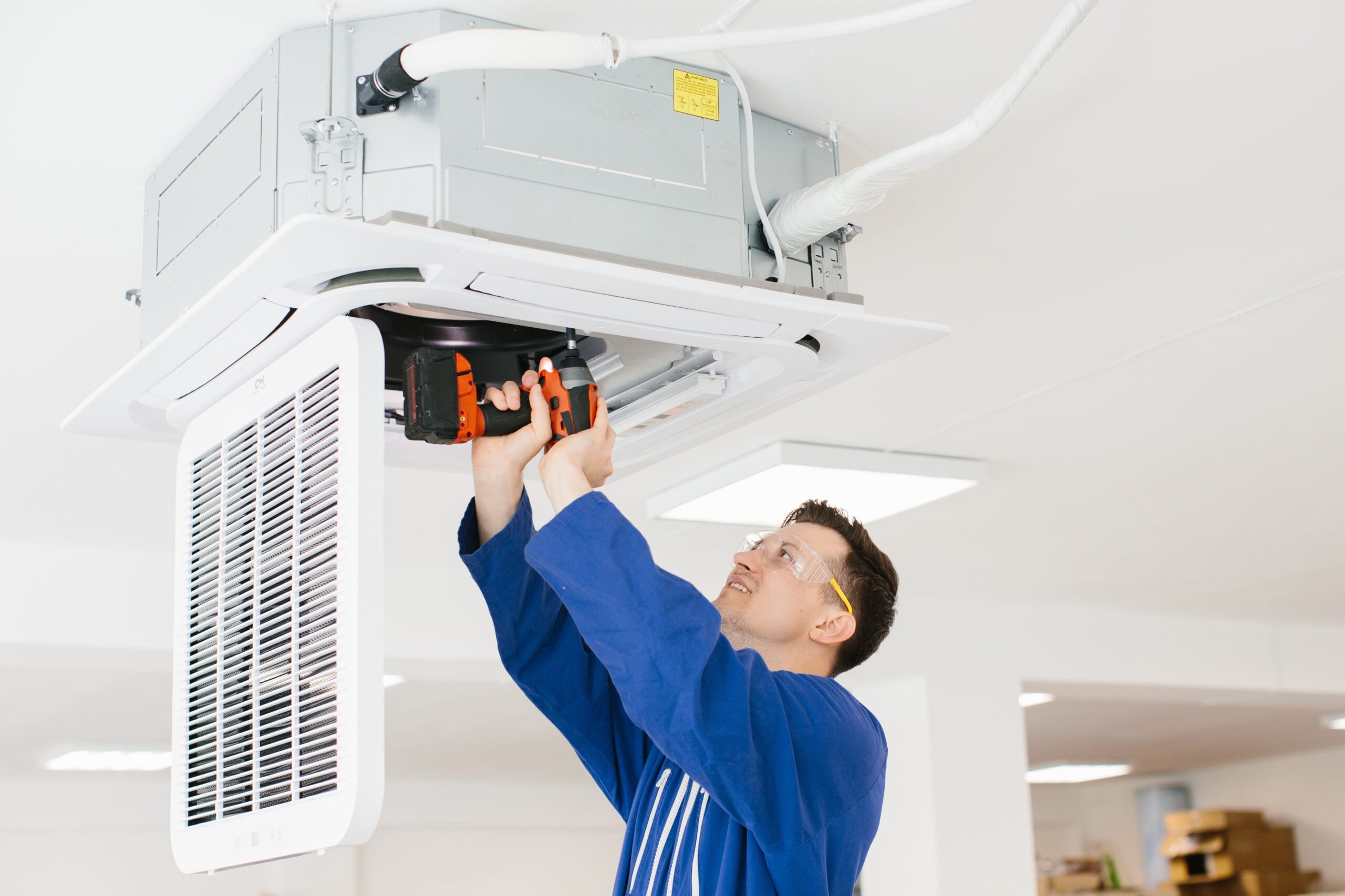 A technician in blue coveralls and safety goggles uses a cordless drill to repair or install a ceiling-mounted air conditioning unit in a brightly lit indoor space.