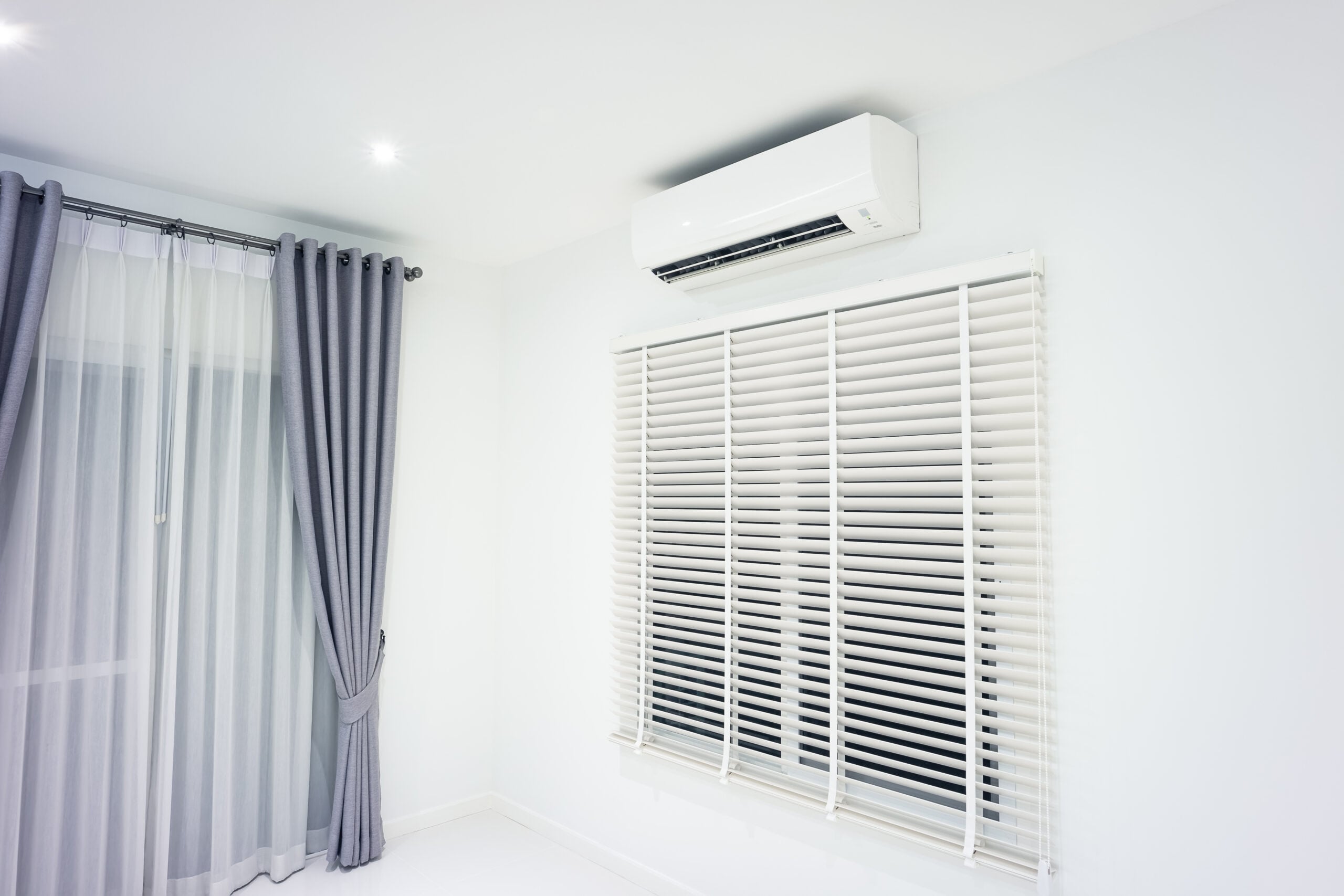 A corner of a room with a window covered by white horizontal blinds, another window with sheer and gray curtains, and a white wall-mounted air conditioner above the blinds.