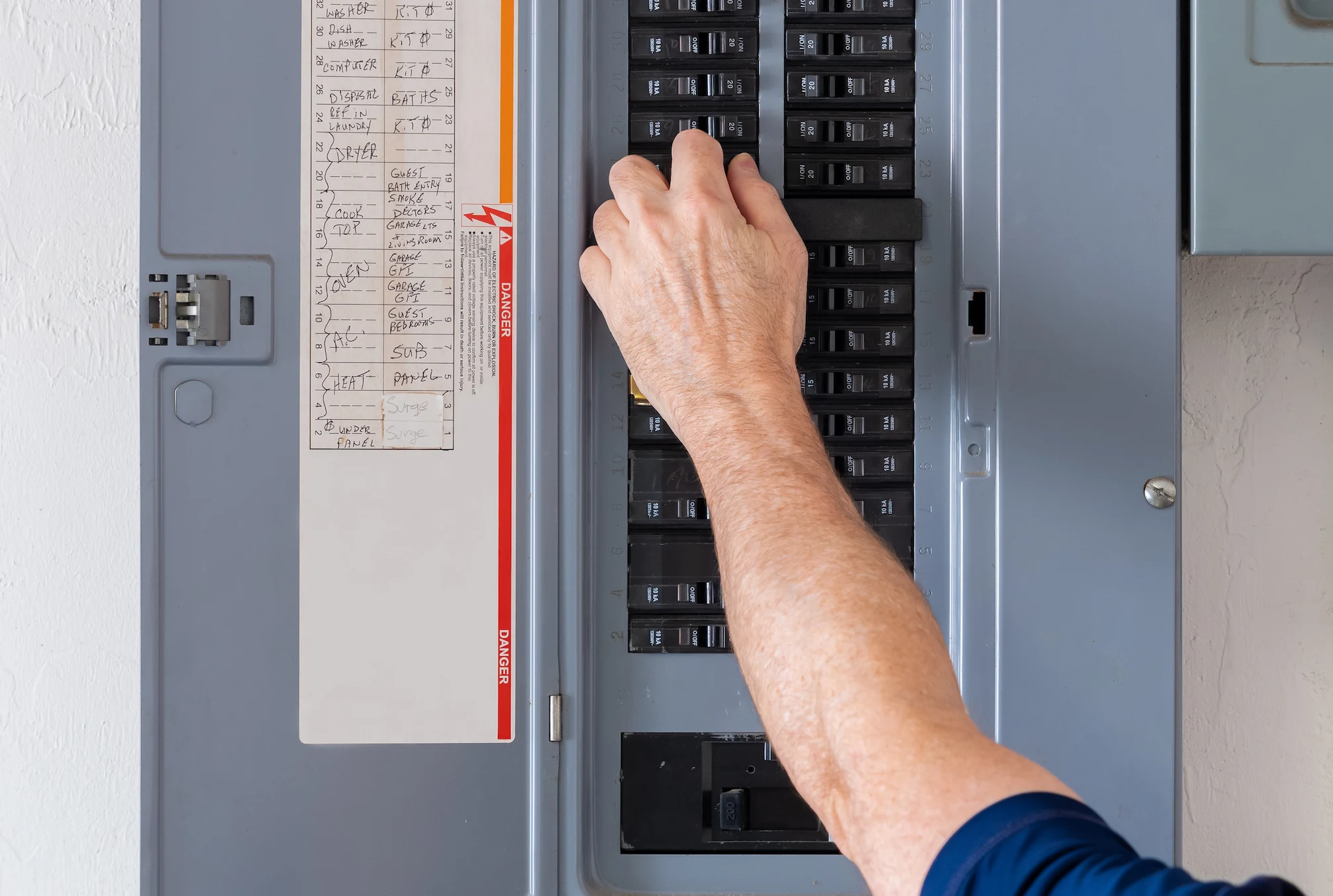 A hand flips a breaker in an electrical panel. The panel has labels for circuits, including WASHER, DRYER, and OVEN.