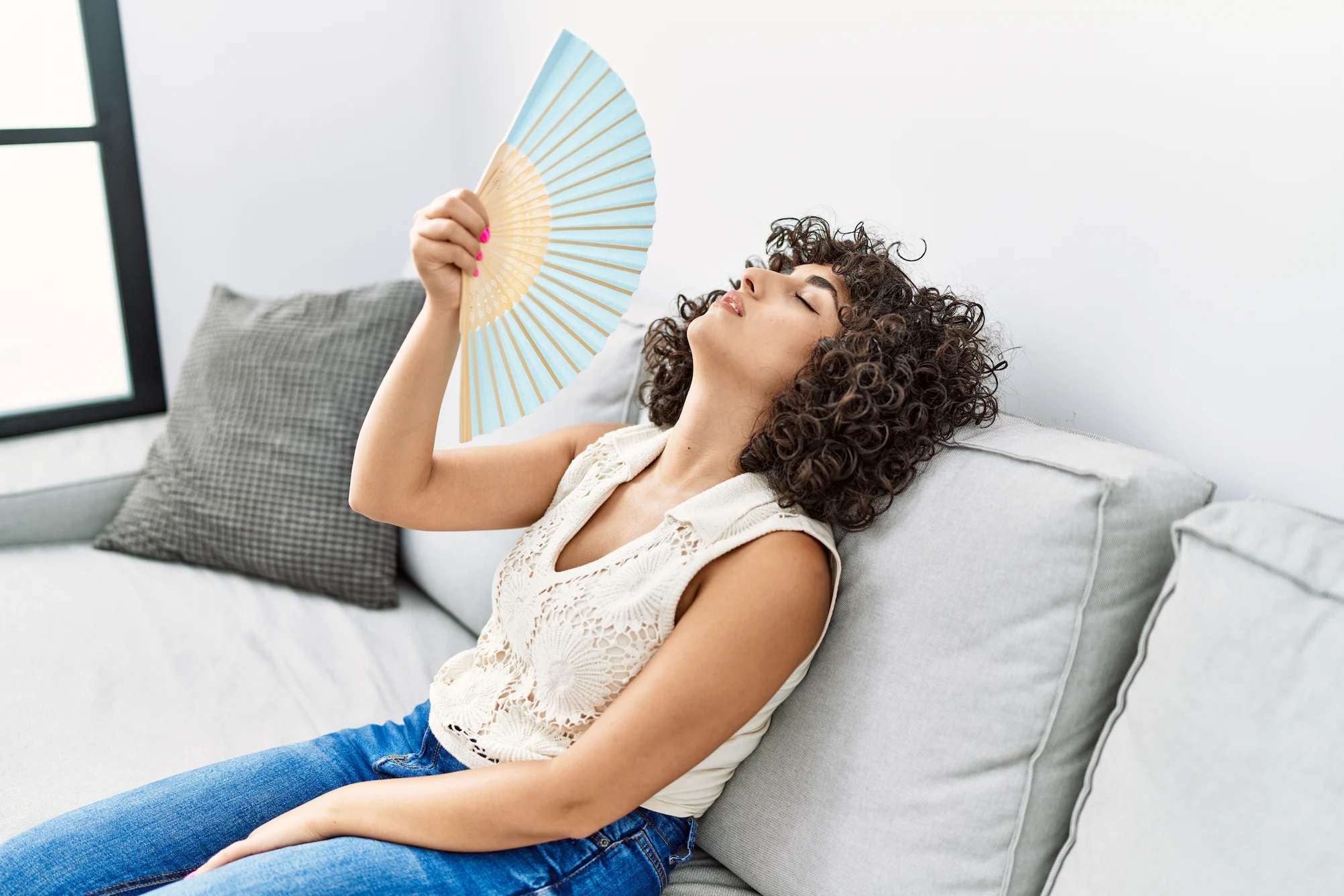 A woman with curly hair fans herself with a blue and yellow hand fan while sitting on a light gray couch.
