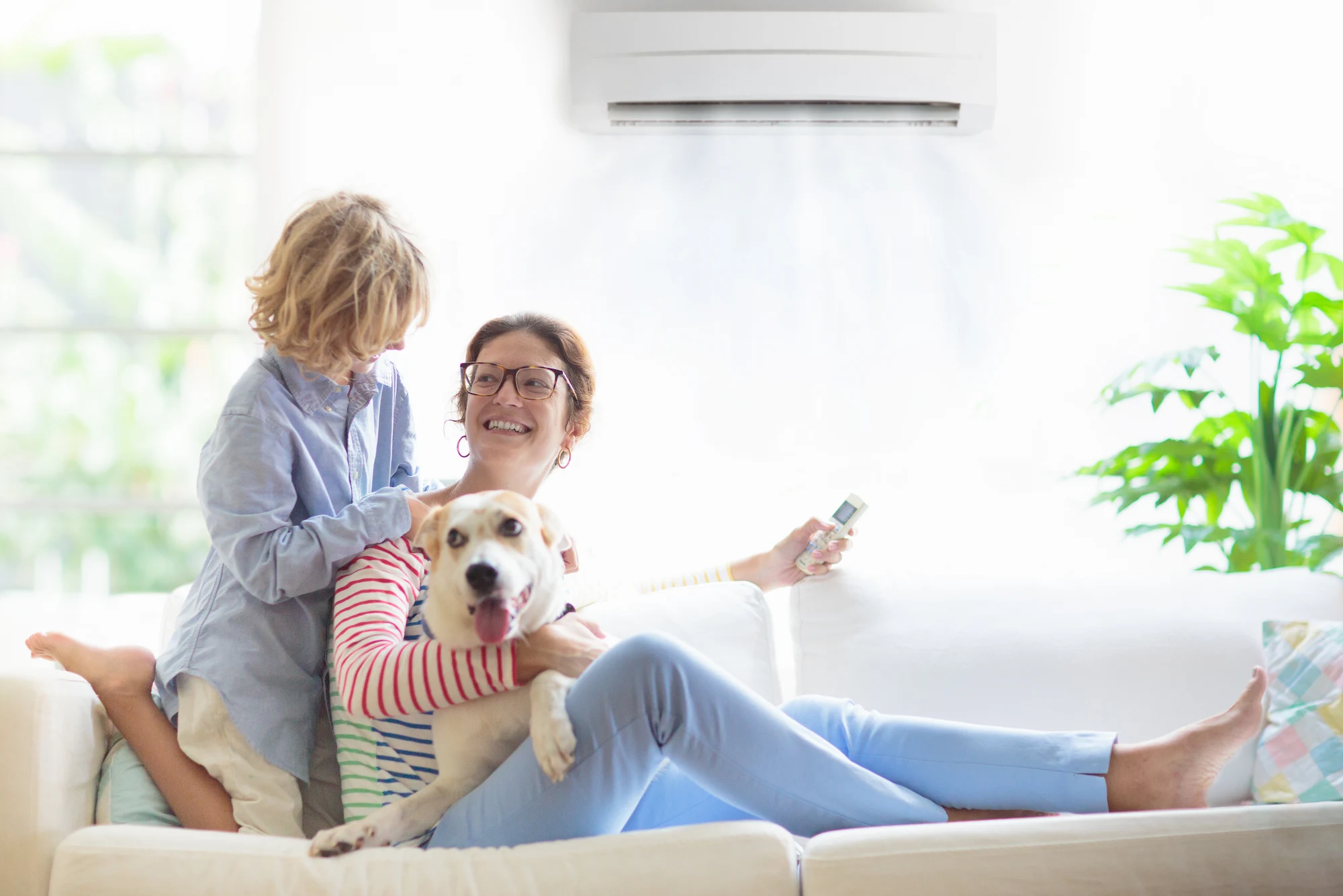 A mother and child sit on a couch with a dog. An air conditioner is mounted on the wall above them.