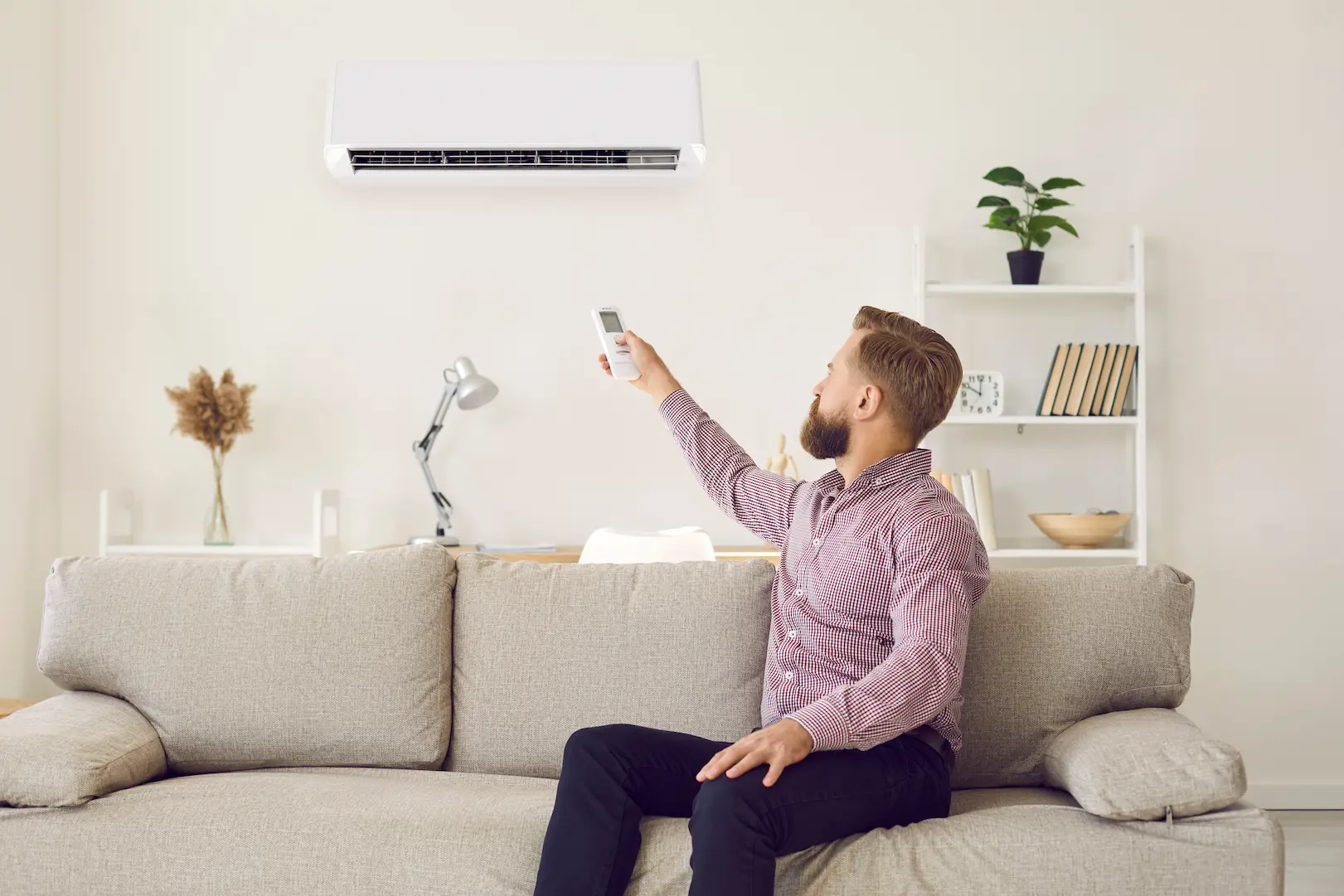 A man with a beard is sitting on a sofa and operating an air conditioner with a remote control.