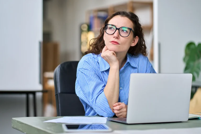 A woman with curly brown hair and glasses sits at a desk with a laptop in front of her, looking up thoughtfully.