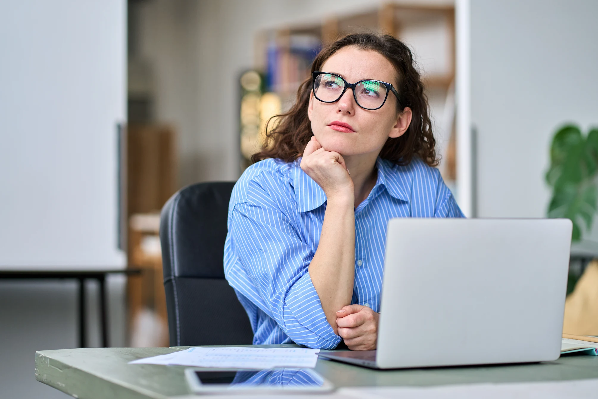 A woman with curly brown hair and glasses sits at a desk with a laptop in front of her, looking up thoughtfully.