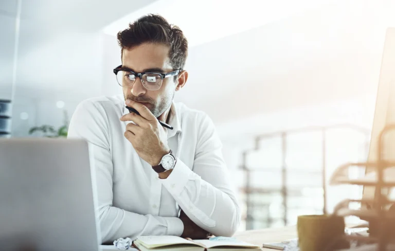 A man wearing glasses and a white shirt sits at a desk, holding a pen to his chin while looking at a laptop.