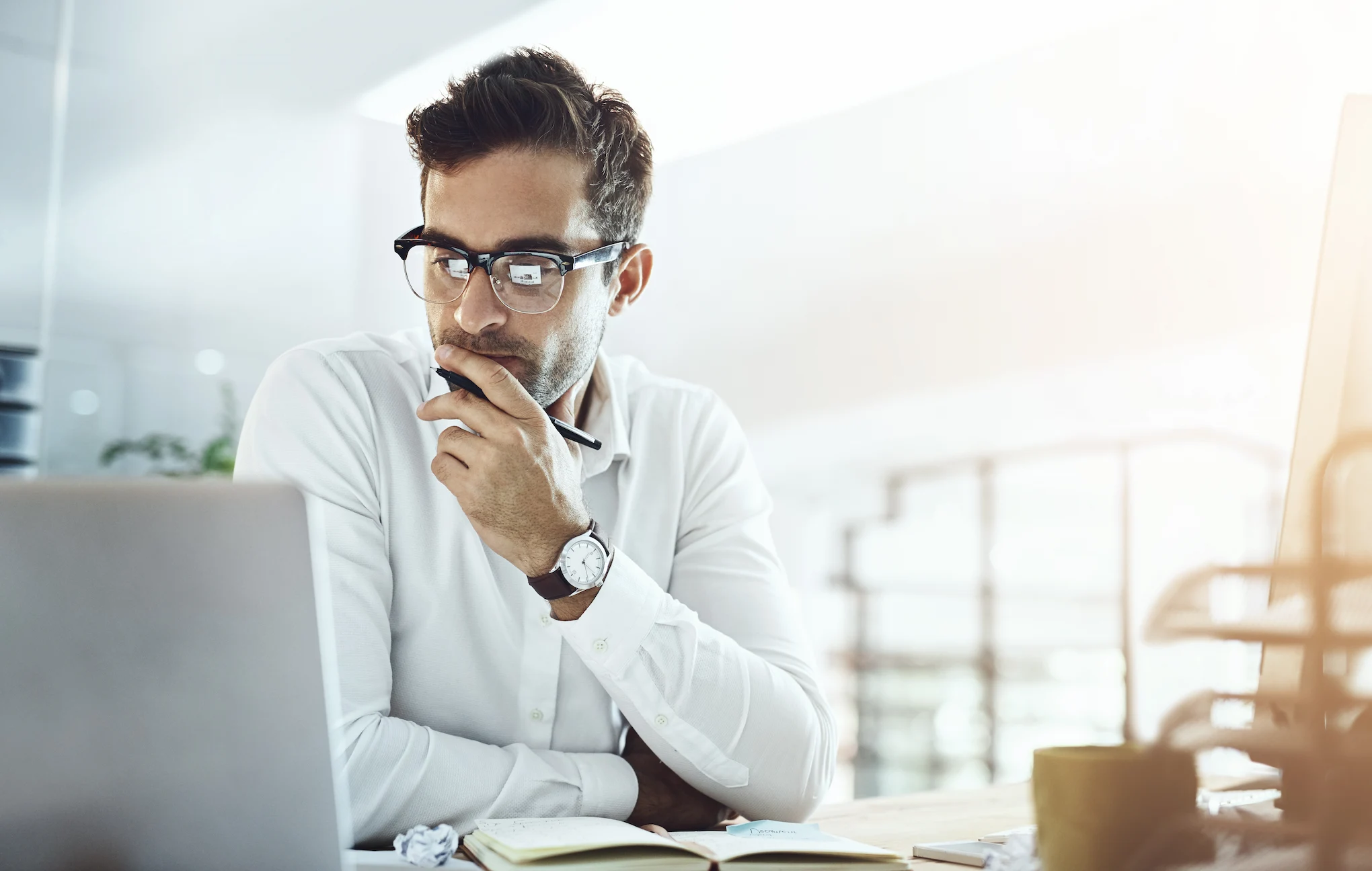 A man wearing glasses and a white shirt sits at a desk, holding a pen to his chin while looking at a laptop.