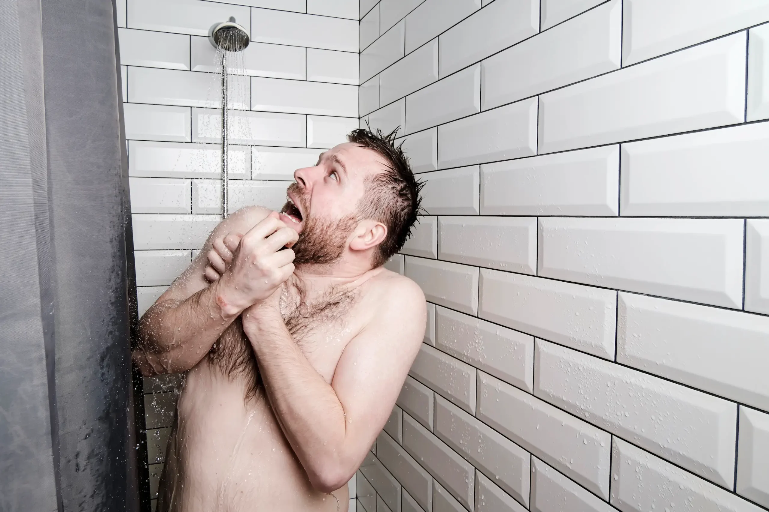 A man standing in a shower with white subway tile walls appears startled and uncomfortable, clutching his hands to his chest and looking upwards toward the shower head.