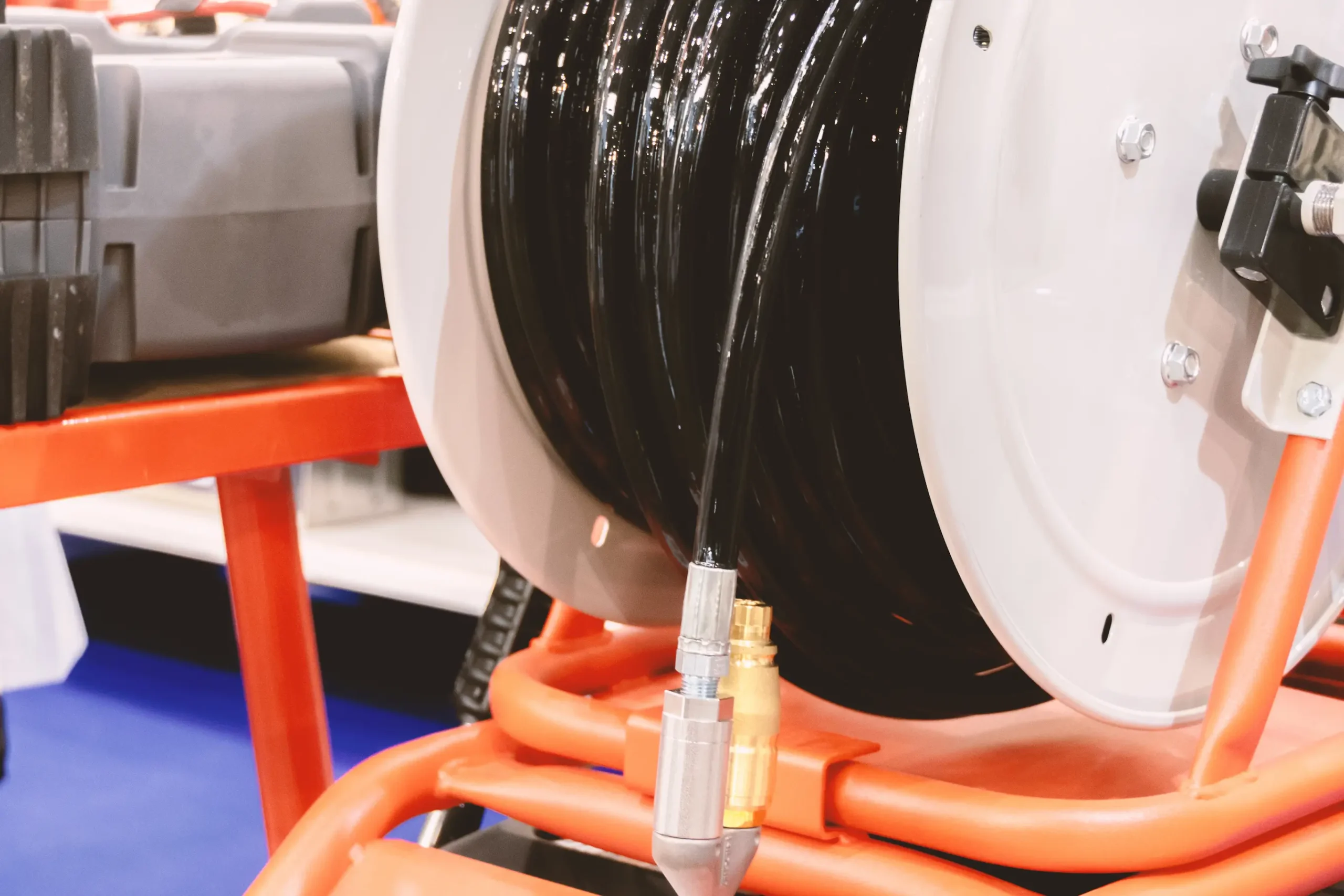 Close-up of a large black hose wound around a reel mounted on an orange metal frame, with a metal hose connector attached to the end; a gray plastic case is in the background.