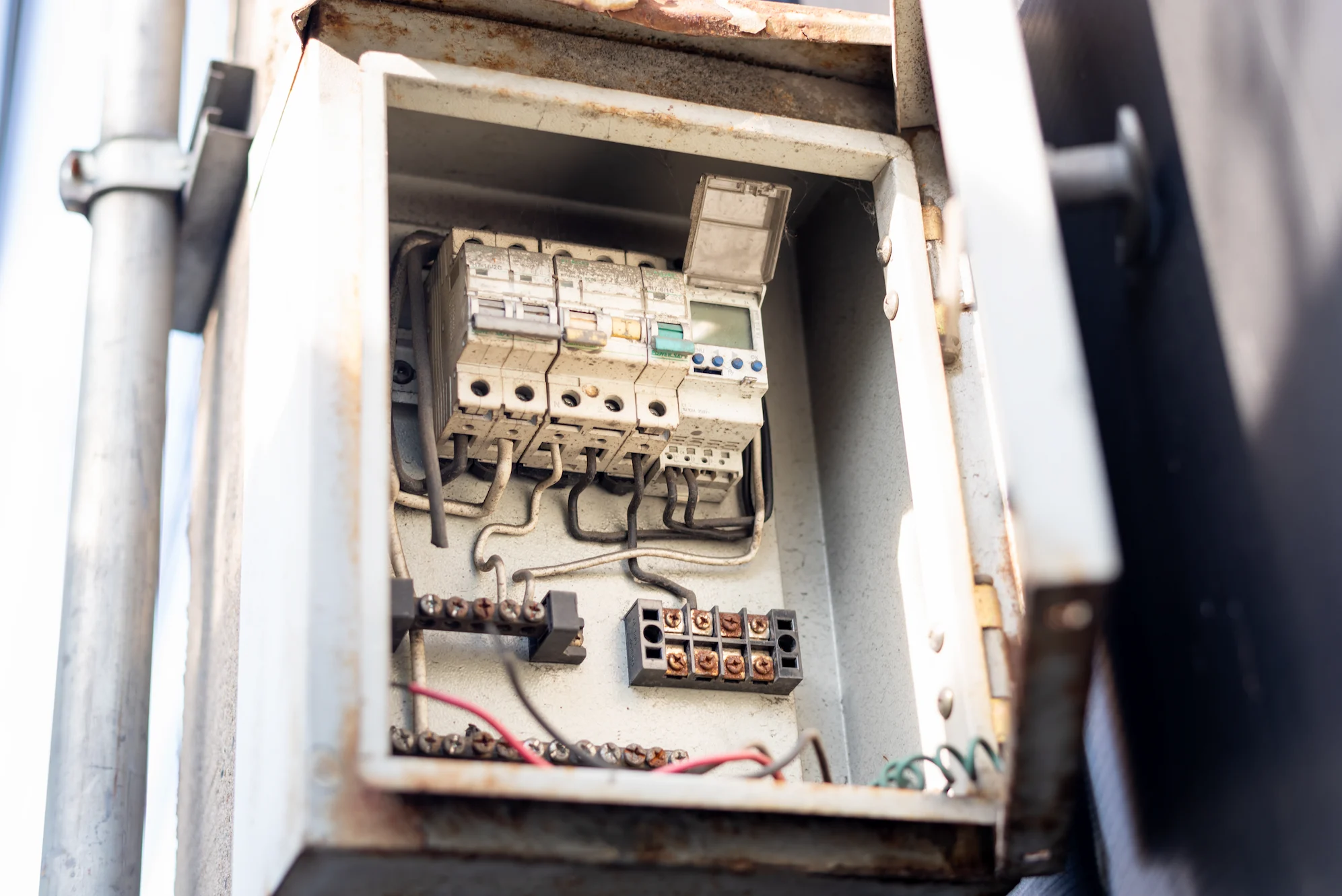 An open metal electrical panel shows circuit breakers, various wires, and connection terminals inside. The panel is slightly rusted and mounted vertically.