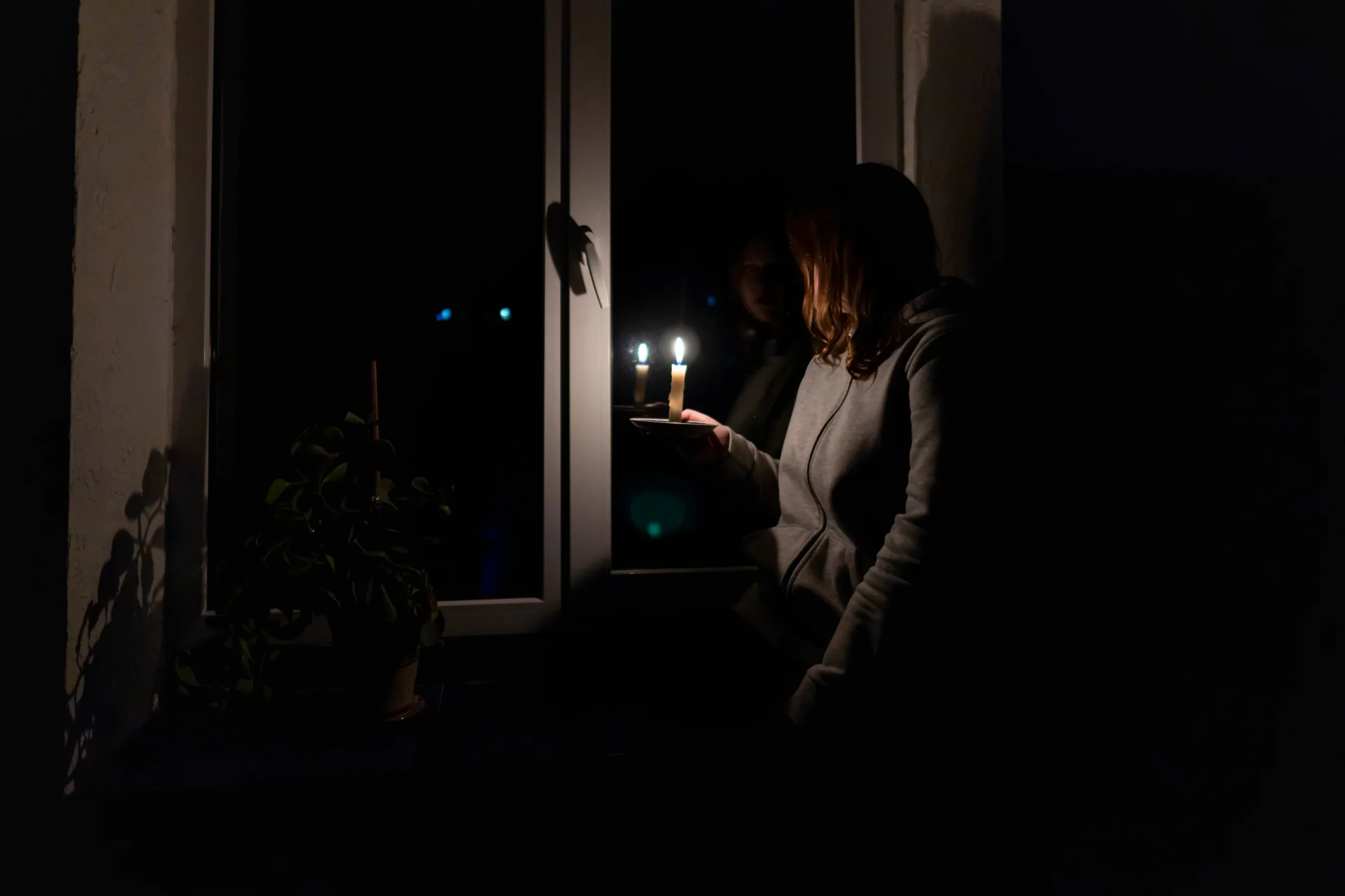 A person in a hoodie sits by a window at night, holding a lit candle. A potted plant is on the windowsill, and the room is mostly dark except for the candlelight.