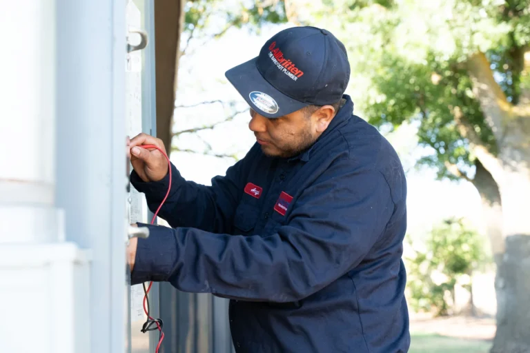 A man wearing a navy blue uniform and cap uses a tool to check electrical wiring on an outdoor panel, with trees and sunlight visible in the background.