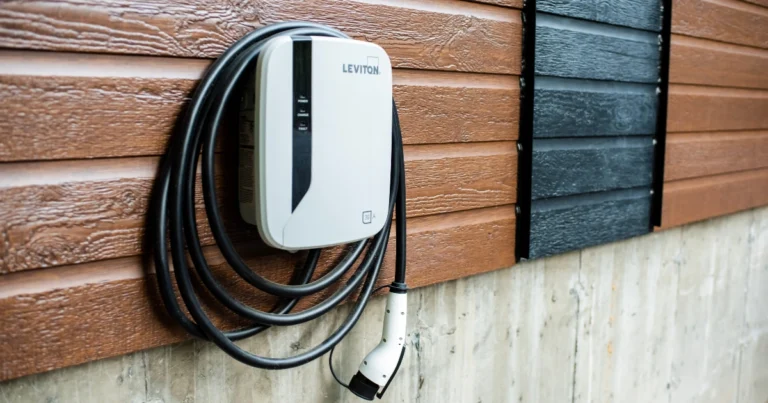 A white Leviton electric vehicle charging station is mounted on a brown wooden wall, with a coiled charging cable and connector hanging neatly below the unit.