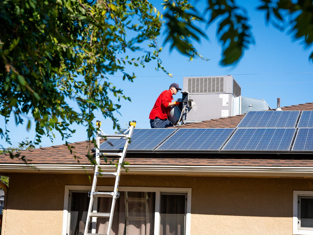 A worker in a red shirt and cap is installing or inspecting solar panels on a house roof, with a ladder leaning against the building. There are trees and HVAC equipment visible.