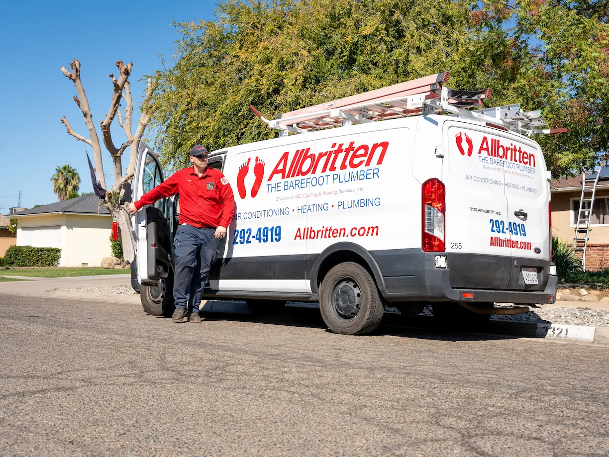 A plumber in a red shirt and cap stands next to a white Allbritten service van with ladders on top, parked on a residential street in front of houses and trees.
