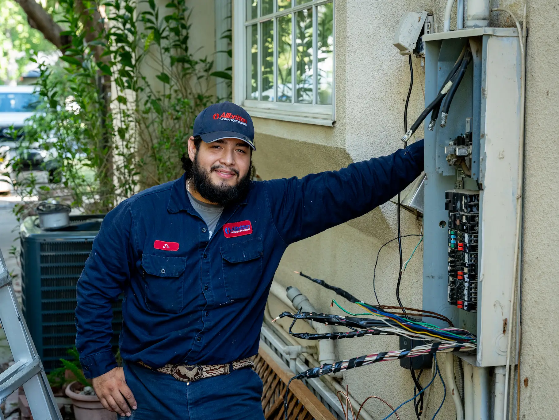 A man in a navy work uniform and cap stands beside an open electrical panel mounted on an exterior wall. He is smiling, with one arm resting on the panel. Wires and equipment are visible inside the panel.