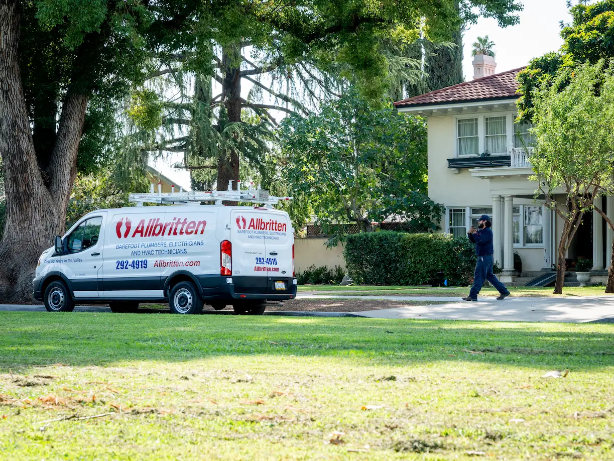A white Allbritten service van is parked on a residential street in front of a house. A worker wearing dark clothing and a cap walks toward the house, carrying equipment. Trees and greenery surround the area.