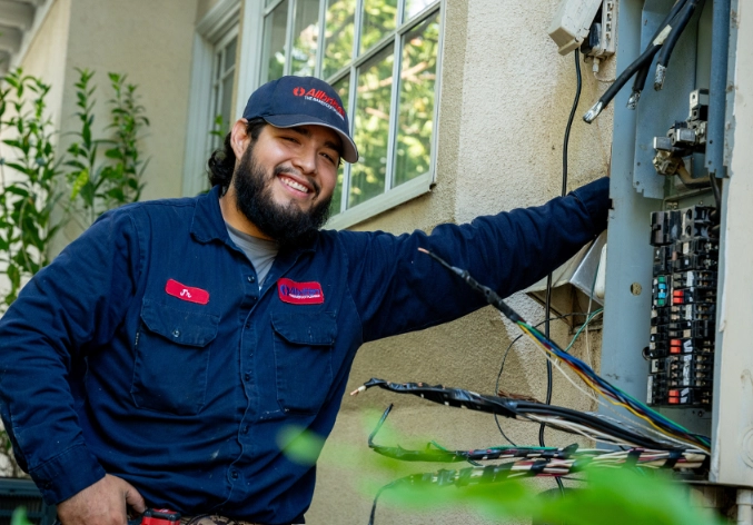 A man in a work uniform and cap stands outdoors by an open electrical panel, smiling at the camera. The panel contains various wires and breakers. There are plants and a window in the background.