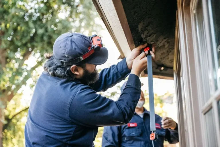 An Allbritten technician wearing a headlamp installs a sensor on the exterior trim of a house while another technician watches nearby.