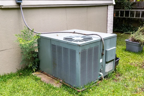 A large, rectangular outdoor air conditioning unit sits on a concrete pad next to a house, surrounded by grass and a few plants, with electrical and drainage connections attached.