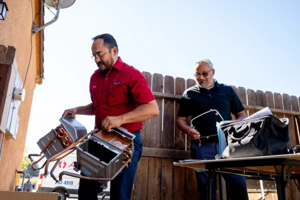 Two men stand outside near a wooden fence, handling metal HVAC parts and tools on a sunny day. One man wears a red shirt and glasses, and the other wears a black shirt and sunglasses. A tool bag is on the table.