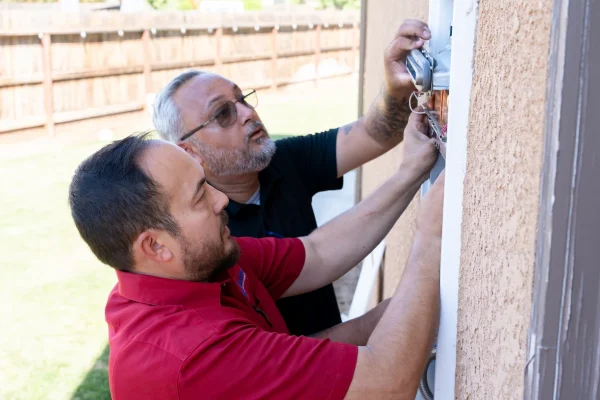 Two men work on an electrical panel mounted on an exterior wall, with one holding the panel cover and the other handling wires. A yard and wooden fence are visible in the background.