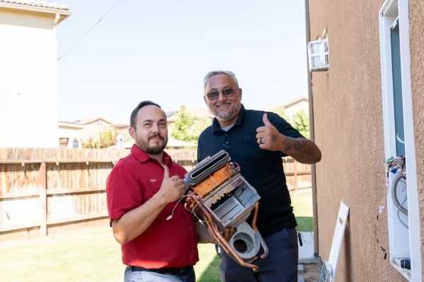 Two men standing outdoors next to a house, one holding HVAC equipment, both giving a thumbs-up gesture and looking at the camera. There is a fenced yard and houses in the background.