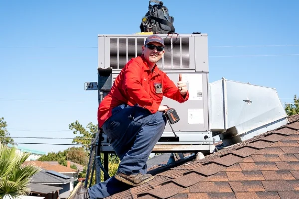 A worker in a red shirt and blue pants kneels on a shingled roof, giving a thumbs up in front of a large HVAC unit. Tools and a black bag are on top of the unit. The sky is clear and blue.