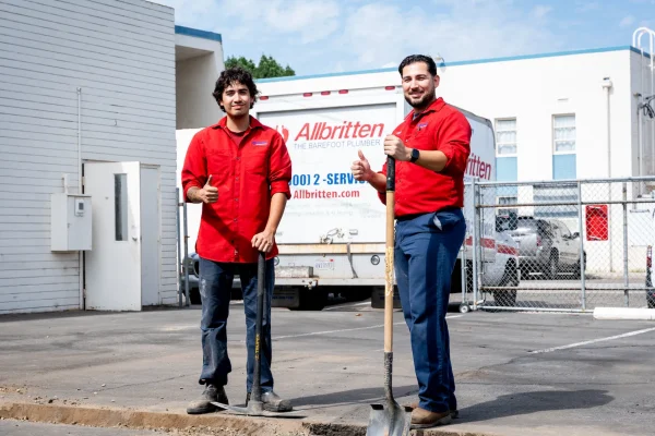 Two men in red shirts and dark pants stand on a paved area next to a dug-up section, each holding a shovel. A white van with Allbritten and a phone number is parked behind them near a building.