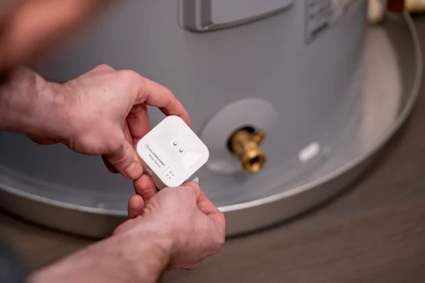 A person holds a small white sensor device near a water heater, positioning it close to a brass valve at the bottom of the appliance.
