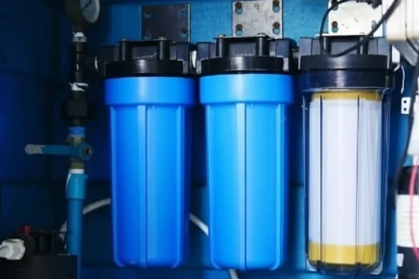 Three water filter cartridges, two solid blue and one transparent with a visible filter inside, are installed in a row as part of a water filtration system against a blue background.